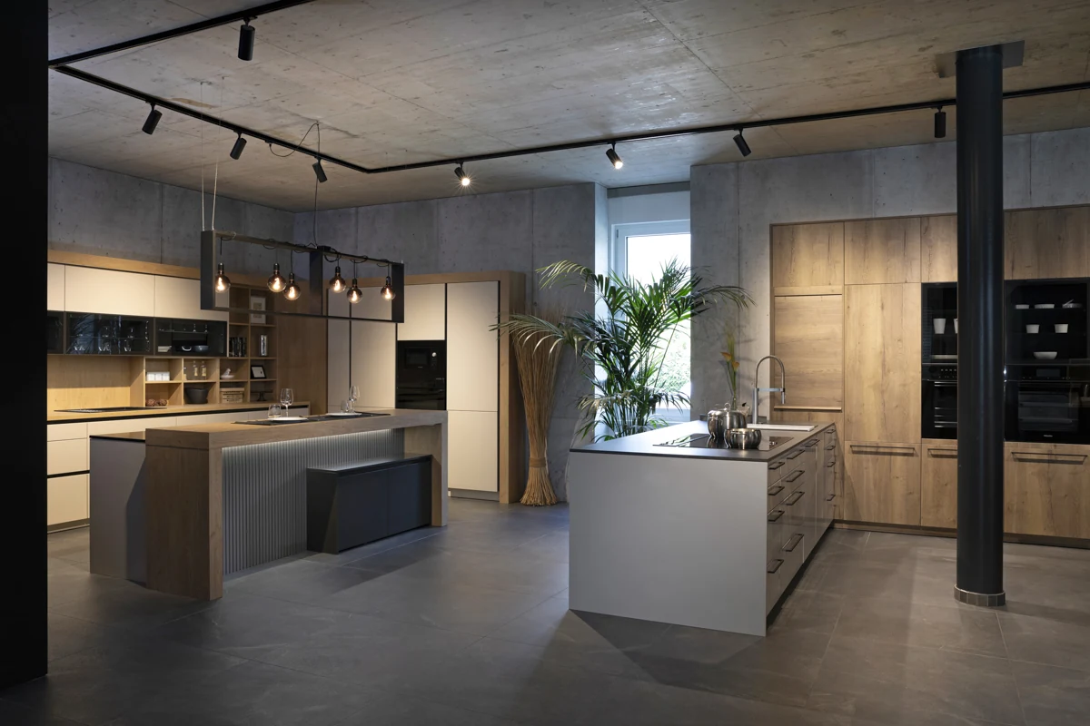 Weiter Blick in unser Küchenstudio mit zwei Kücheninseln, Holzfronten und warmen Pendelleuchten